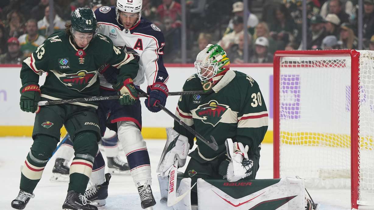 Minnesota Wild goaltender Jesper Wallstedt (30) and defenseman Quinn Hughes (43) defend against a shot as Columbus Blue Jackets center Sean Monahan (23) pressures them during the first period of an NHL hockey game, Sunday, Oct. 18, 2026, in St. Paul, Minn.