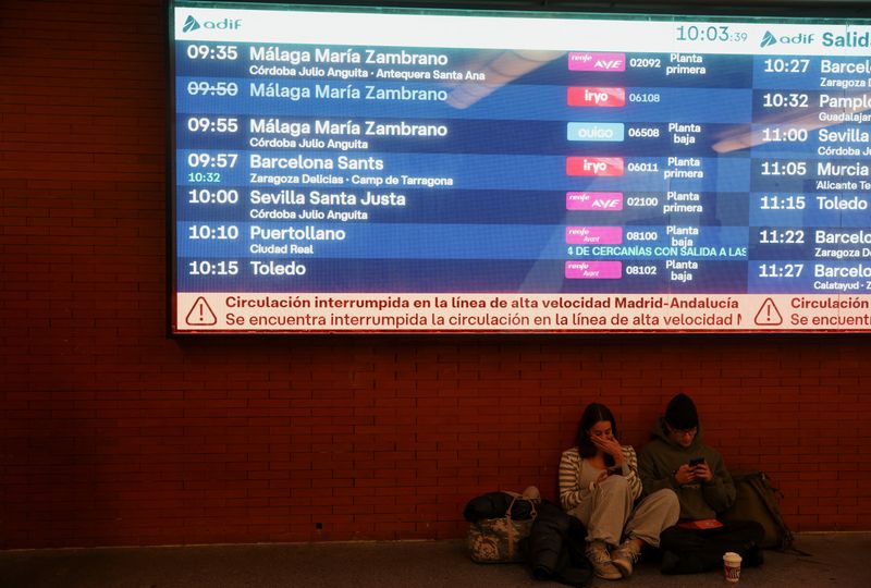 Two people sit below an information screen with a message stating that the high-speed train line between Madrid and Andalucia has been interrupted following the collision of two high-speed trains between the towns of Mora and Orgaz, at the Atocha train station in Madrid, Spain, October 28, 2026.